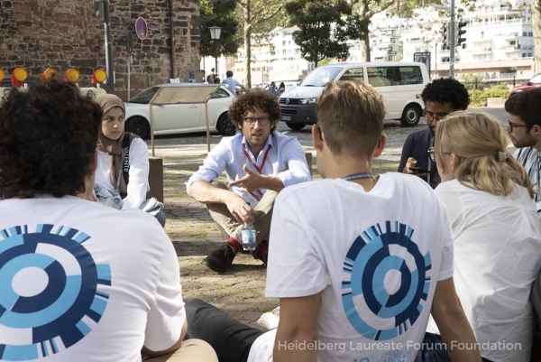 A man with light skin and curly dark hair who is wearing a red lanyard sits cross-legged on the ground in an apparent circle of students, who are wearing white T-shirts that have a blue logo.