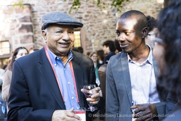 An older man of Indian descent in a cap stands next to a young Black man, both wearing open-collared shirts with blazers.