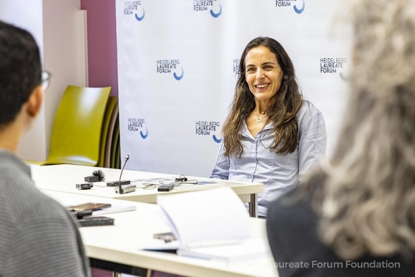 A woman with light skin and long brown hair smiling at a press conference while journalists whose faces are unseen ask questions.