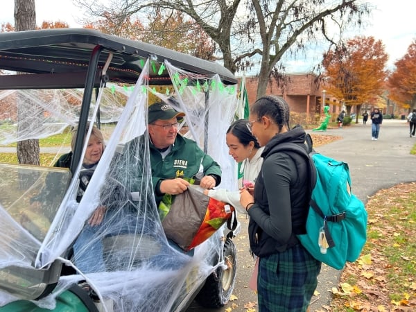 President Chuck Seifert and his wife sit inside a green golf cart covered in fake cobwebs. Seifert holds open a bag of Halloween candy to two students on campus.