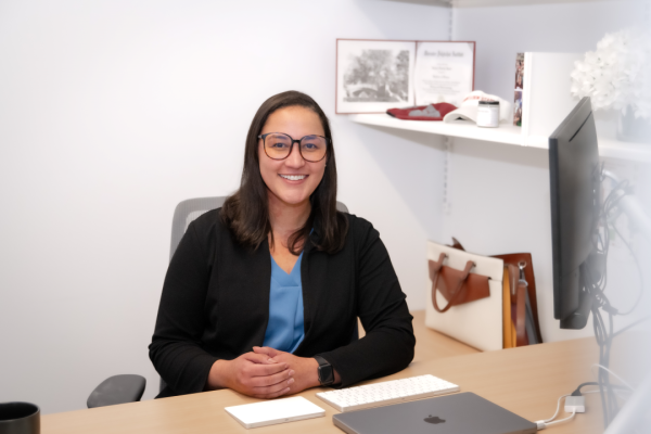 Rachael Heard, who has dark hair and glasses, sits behind her desk wearing a black blazer and blue blouse.