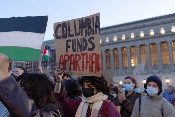 A student in a scarf and mask hold a sign saying, "Columbia Funds Apartheid."
