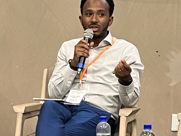 A dark-skinned man with black hair and a beard, wearing a white shirt and jeans and an orange lanyard around his neck, speaks into a microphone while sitting in a chair.