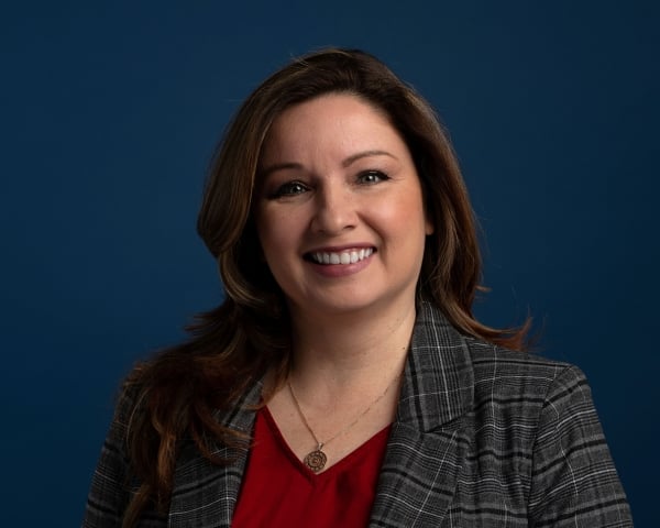 Marilyn Jones, a light-skinned woman with shoulder-length brown hair, smiles in a red blouse and black plaid blazer