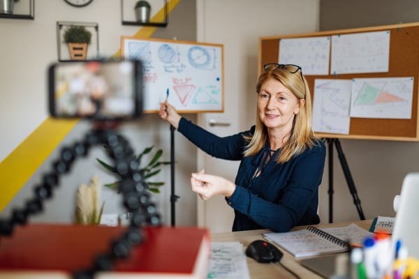 A woman points toward a whiteboard while a cellphone on a tripod films her