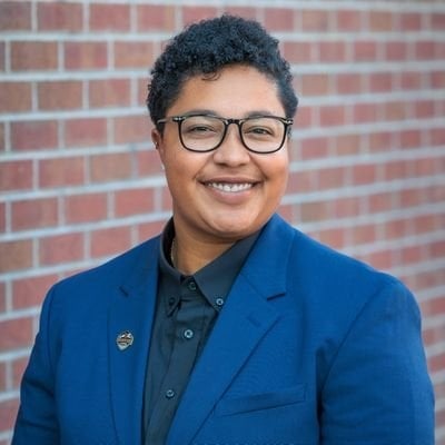 Ashlie Prioleau smiles for a headshot wearing glasses, a blue suit jacket and gray collared shirt