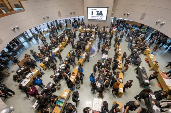 An aerial view of a large multipurpose room at Bryant University filed with yellow and black tables featuring prototypes of student ideas.