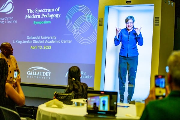 A digital woman appears in a box. She is holding up her hands showcasing sign language and is in front of a room filled with people