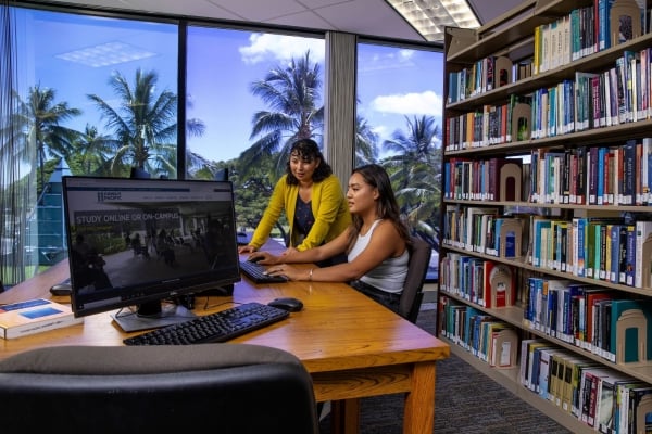 A female student and teacher are in a library looking at a computer. There are palm trees in a window behind the woman and student. 