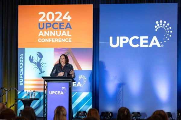 A woman is on a stage standing behind a podium. There is a background behind him with "2024 UPCEA" emblazoned on the blue and orange tarp. 
