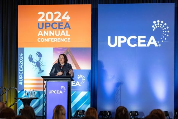 A woman is on a stage standing behind a podium. There is a background behind him with "2024 UPCEA" emblazoned on the blue and orange tarp.