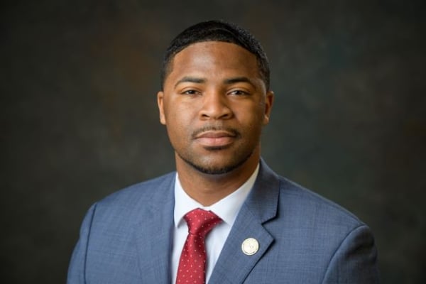 Gilbert Rogers poses for a headshot in a gray suit jacket and red tie, with a gold pin on his lapel