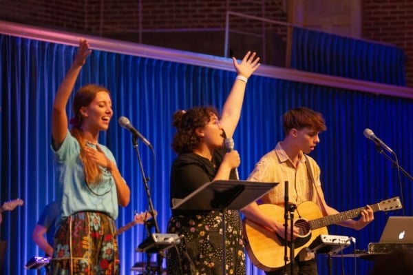 Moody Bible Institute students sing in front of microphones with raised hands, one playing guitar.