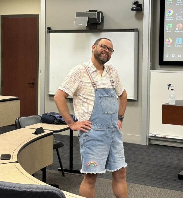 A man in short coveralls with a rainbow patch and a button down shirt