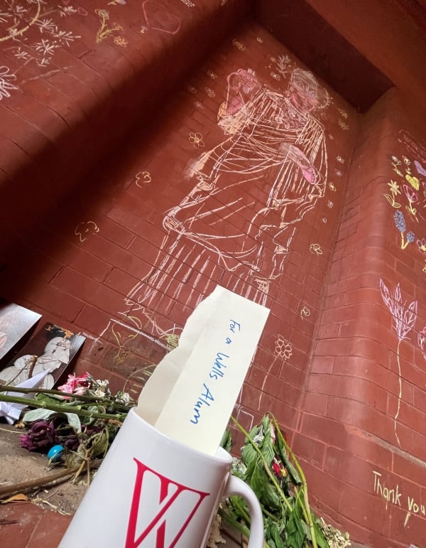 A photo of a makeshift memorial at Wells College, with flowers and a mug with a note tucked in that reads "For a Wells alum."