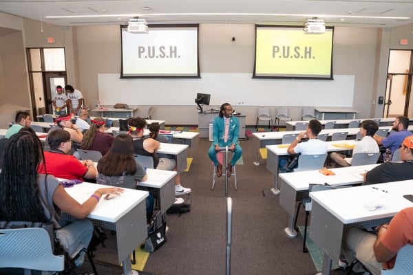 A professor sits in a chair in the middle of rows of students in a classroom, with the acronym P.U.S.H. on the two screens behind him. 