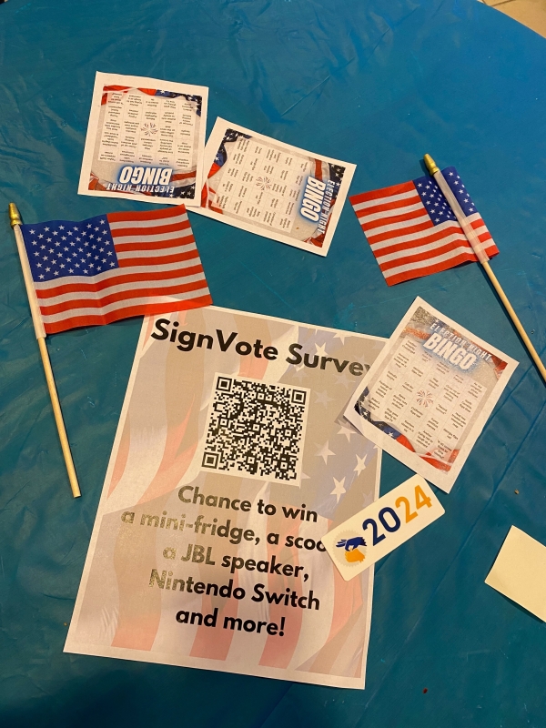 Two American flags, three bingo sheets, a survey QR code and a sticker with "2024" printed on it are displayed on a blue table cloth