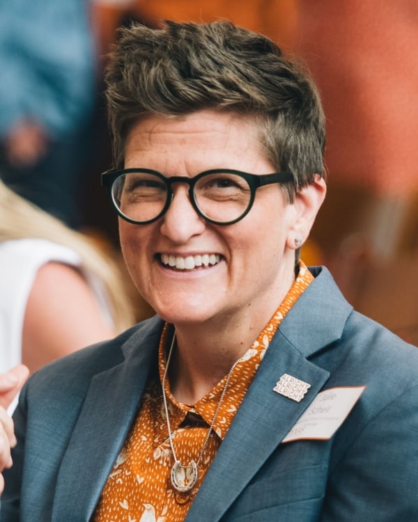 Julie Schell smiles for a headshot in a gray blazer and orange button-up shirt. Julie wears black-framed glasses.