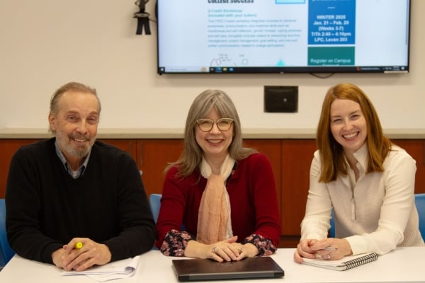 Three faculty members sit at a table with their hands in front of them. In the background is a large TV with the syllabus for the course.