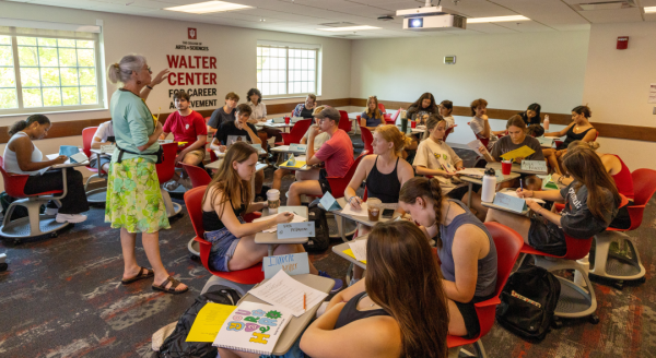 A group of students work at desks in a classroom, while an instructor gives directions at the left side of the frame.