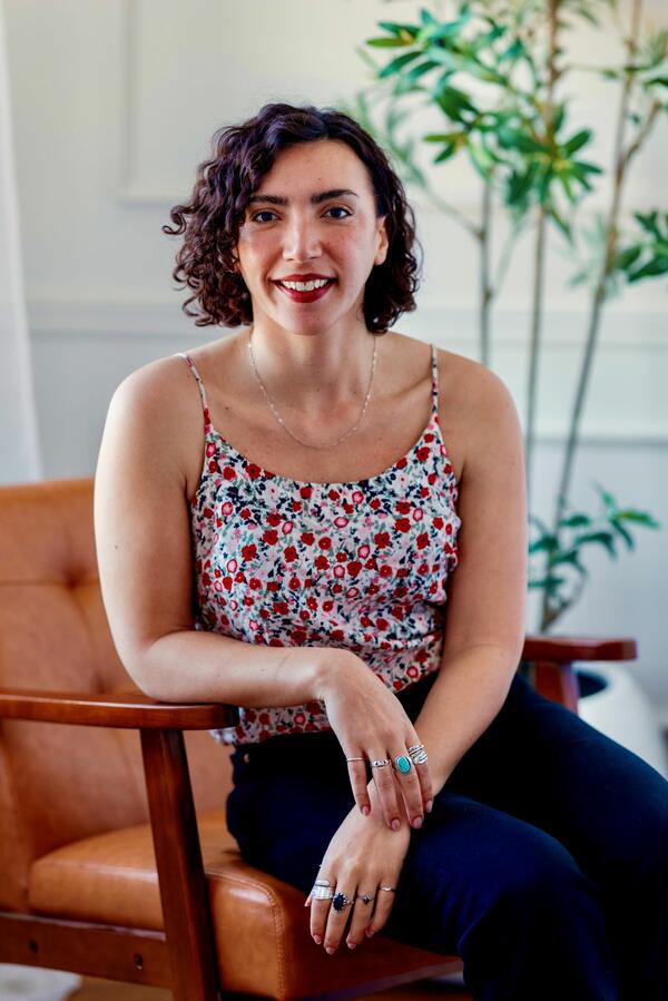 Elyssa Smith smiles for a photo wearing a floral tank top and dark pants sitting in a chair indoors.