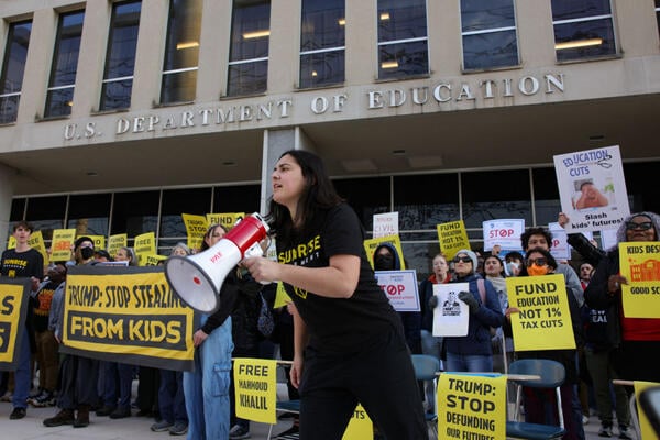 A person in a black shirt holds a megaphone in front of the Education Department, surrounded by people holding yellow signs that say "Trump: Stop Stealing From Kids"