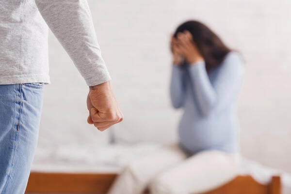 A photo of a pregnant woman with her face in her hands, seemingly scared of her partner, whose body can only be partially seen in the frame.