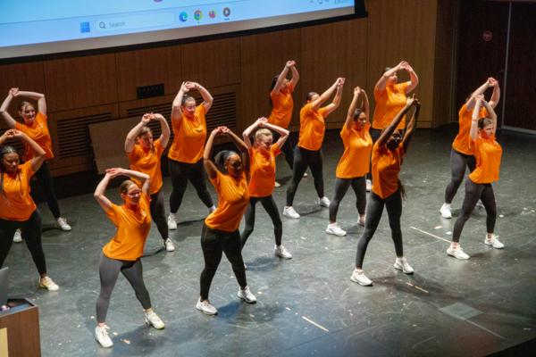 A group of students wearing black leggings, white sneakers and orange shirts perform a dance on stage