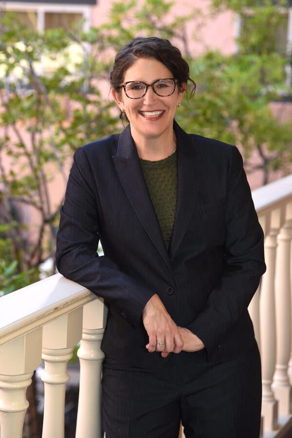 Michelle Deutchman, a light-skinned woman wearing glasses and a dark suit over a dark green top.