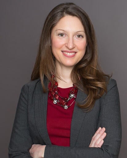 Headshot of a light-skinned woman with shoulder-length brown hair wearing a red blouse under a gray blazer with a statement necklace.