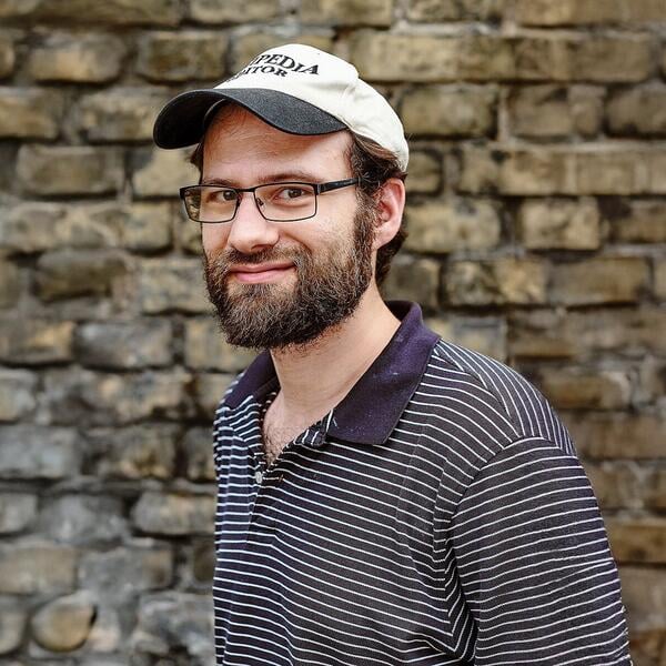 Headshot of a light-skinned man with glasses and a brown beard wearing a striped polo shirt and a baseball cap