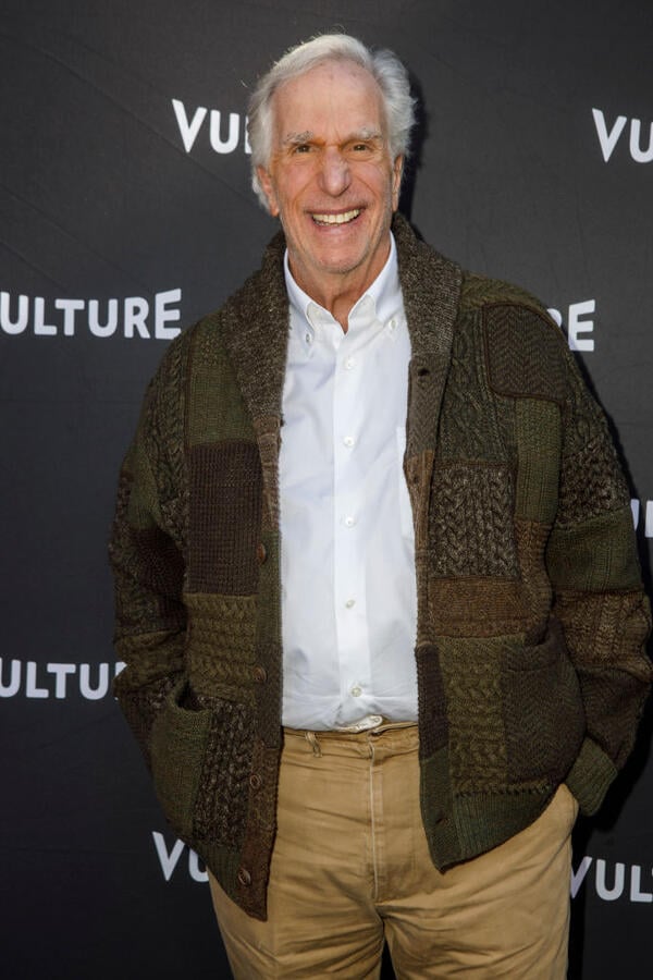 Henry Winkler, an older man with white hair, smiles at the camera. He is wearing a white button-up shirt and a brown jacket.