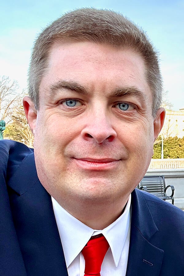 Headshot of S. Daniel Carter, a light-skinned man with short brown hair and very blue eyes, wearing a blue suit, white shirt, and red tie.