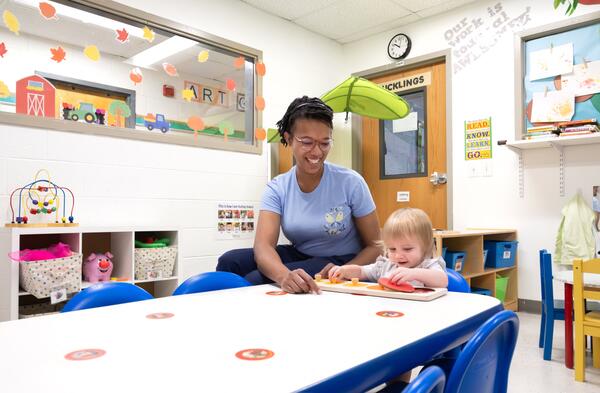 Smiling woman sits next to a young child in a daycare center.