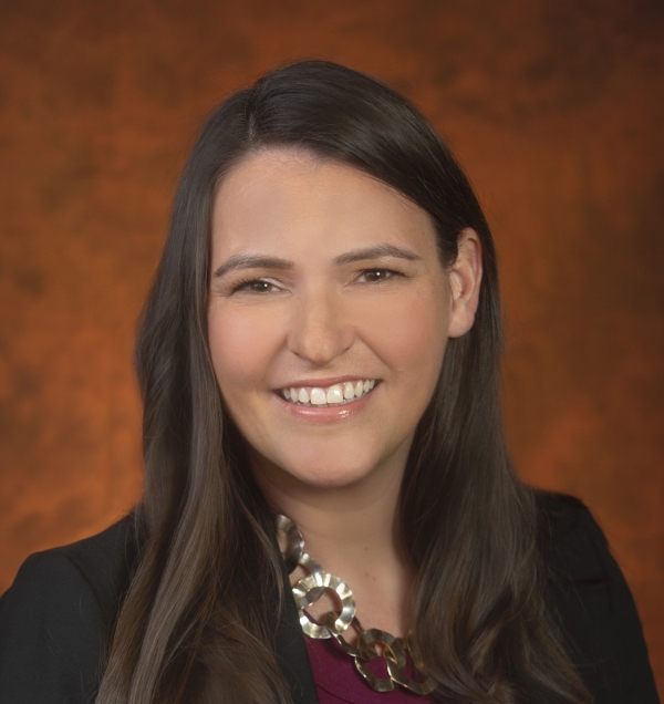 Angela Lauer Chong smiles for a headshot wearing a black blazer and silver necklace in front of an orange backdrop .