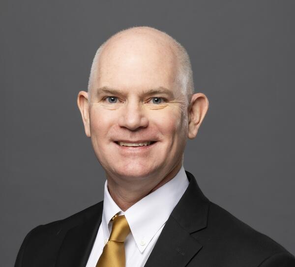 A headshot of Thomas Cavanagh, a light-skinned man with very short gray hair, wearing a dark suit over a white shirt and gold tie.