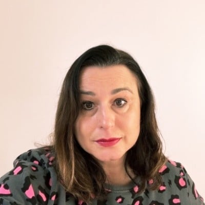 Clare Rawlins, a light-skinned woman with dark hair wearing a patterned top and bright lipstick.