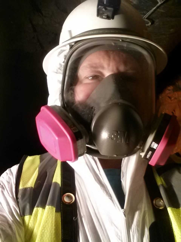 A light-skinned man wearing protective gear, including a high-visibility vest, a respirator over his face, and a helmet.