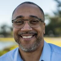 Headshot of David Overton, a man with medium brown skin and a mustache and beard, wearing glasses and a blue shirt.