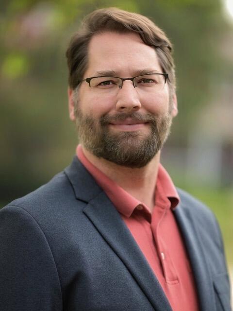 Drew Lurker, a light-skinned white man with brown hair and a bears, wearing glasses that are frameless on the bottom of the lenses.
