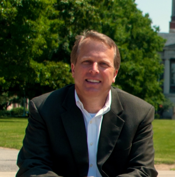 Eric Fredericksen, a light-skinned man with sandy blond hair wearing a black blazer over an open-collared white button-down shirt, sitting in front of a lawn and some trees.