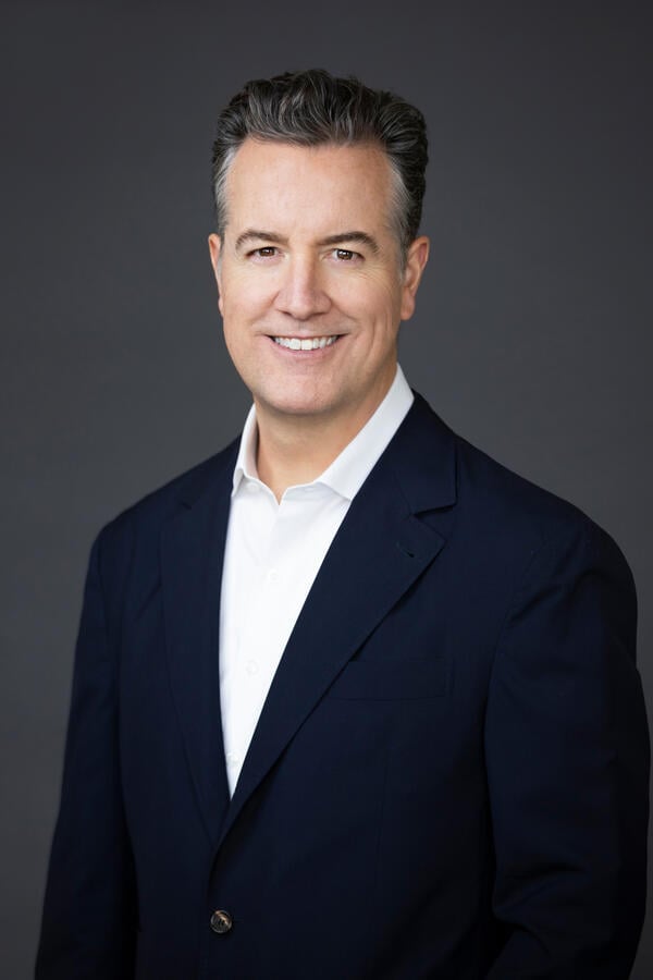 A headshot of Greg Hart, a light-skinned man with dark hair, wearing a dark suit jacket over a white collared shirt open at the neck.