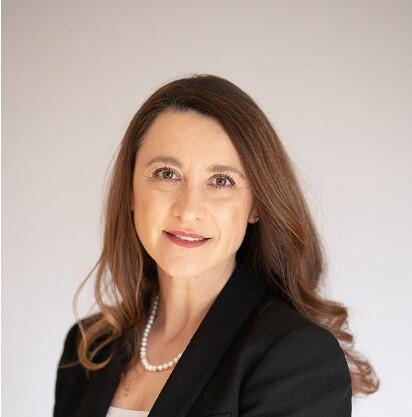 Cabeça de uma mulher de pele clara, com longos cabelos castanhos, vestindo um blazer preto e um colar de pérolas.