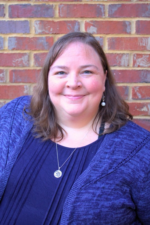 Liz Norell, a light-skinned woman with shoulder-length brown hair, wearing a purple-blue sweater set and a silver pendant necklace.