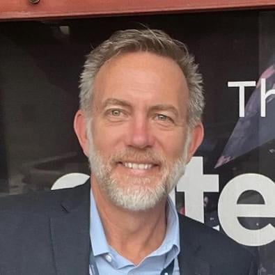 Nathan Greeno, a white man with a gray beard, wearing a blue collared shirt under a dark blazer.