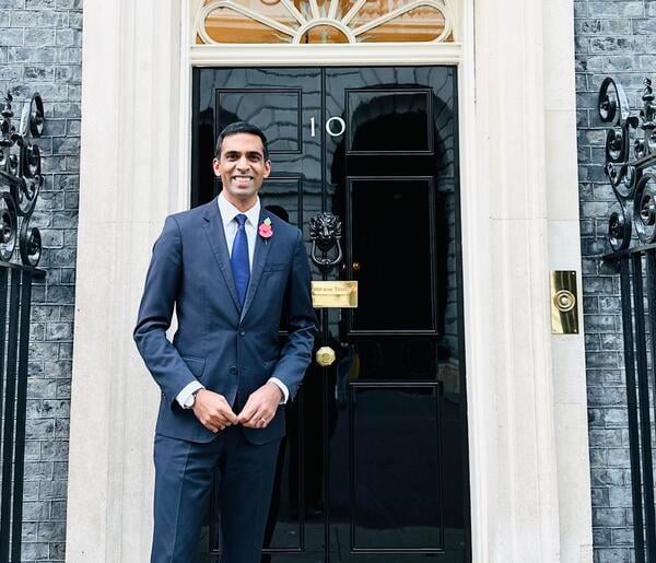 A man with light brown skin and dark hair, wearing a blue suit, stands in front of the recognizable black door of No. 10 Downing Street.