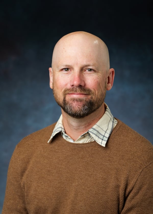 A light-skinned man with a bald head and a beard, wearing a brown sweater over a checked collared shirt.