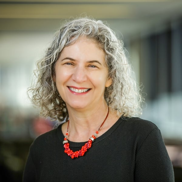 Rebecca Stein, a light-skinned woman with curly gray hair, wearing red lipstick and a black top with a red necklace.
