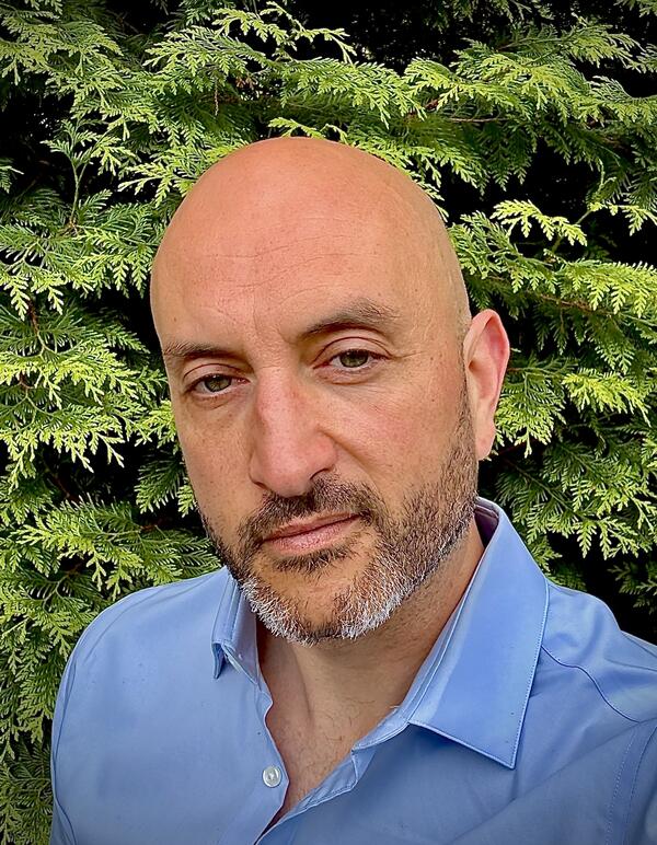 TJ Kalaitzidis, a man with tan skin and a bald head and dark beard with some gray, wearing a blue collared shirt with the top two buttons open.