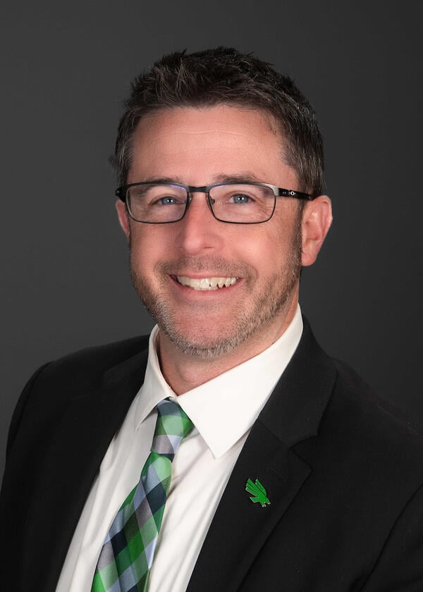 A light-skinned man with short dark hair wearing a black suit coat, white shirt, and green and gray checkered tie.